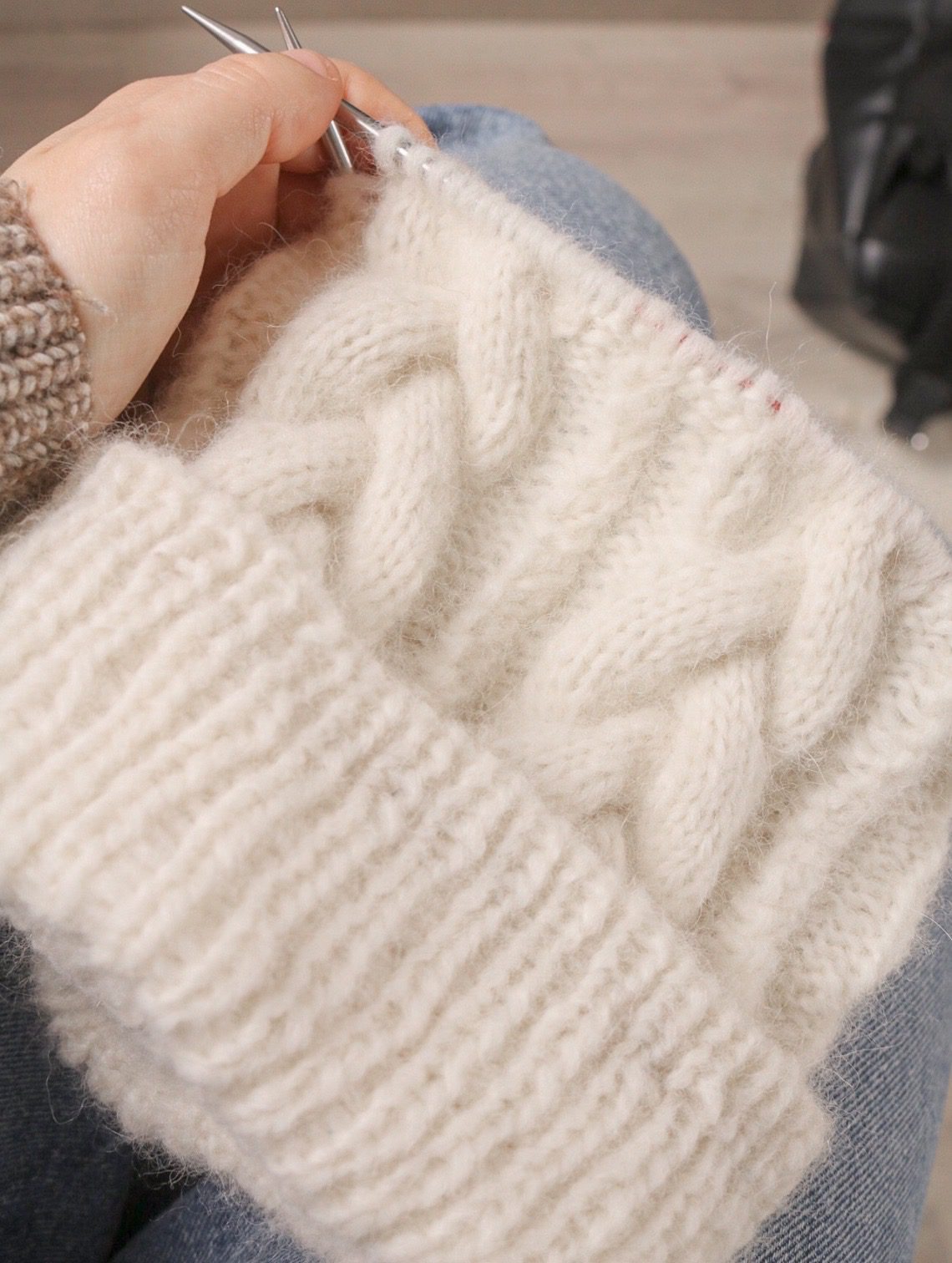 Hands knitting a cream cabled hat on circular needles in DROPS Air, the blown yarn showing a soft halo around each stitch