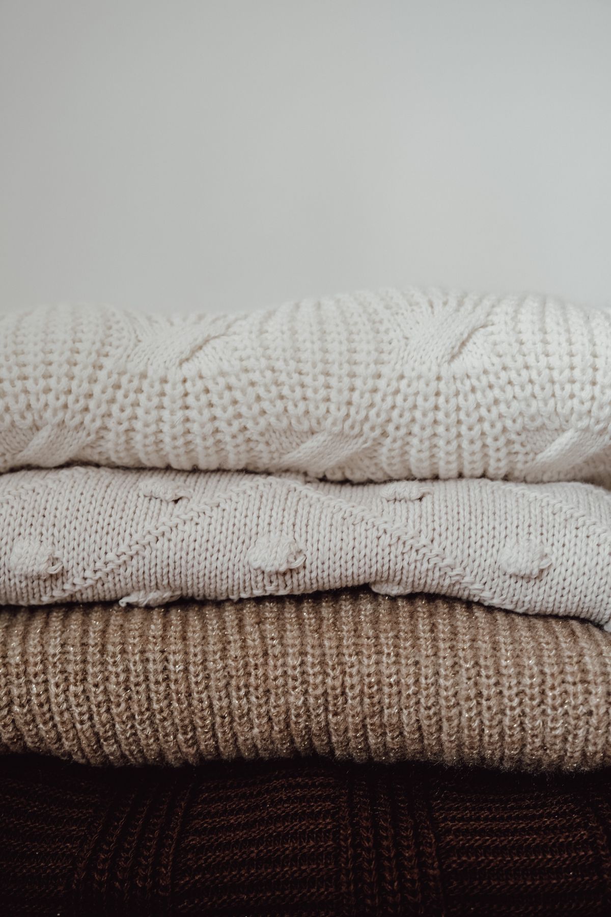 A stack of folded knitted sweaters in cream and warm brown, the kind of second-hand jumpers a crocheter would unravel to reclaim the yarn