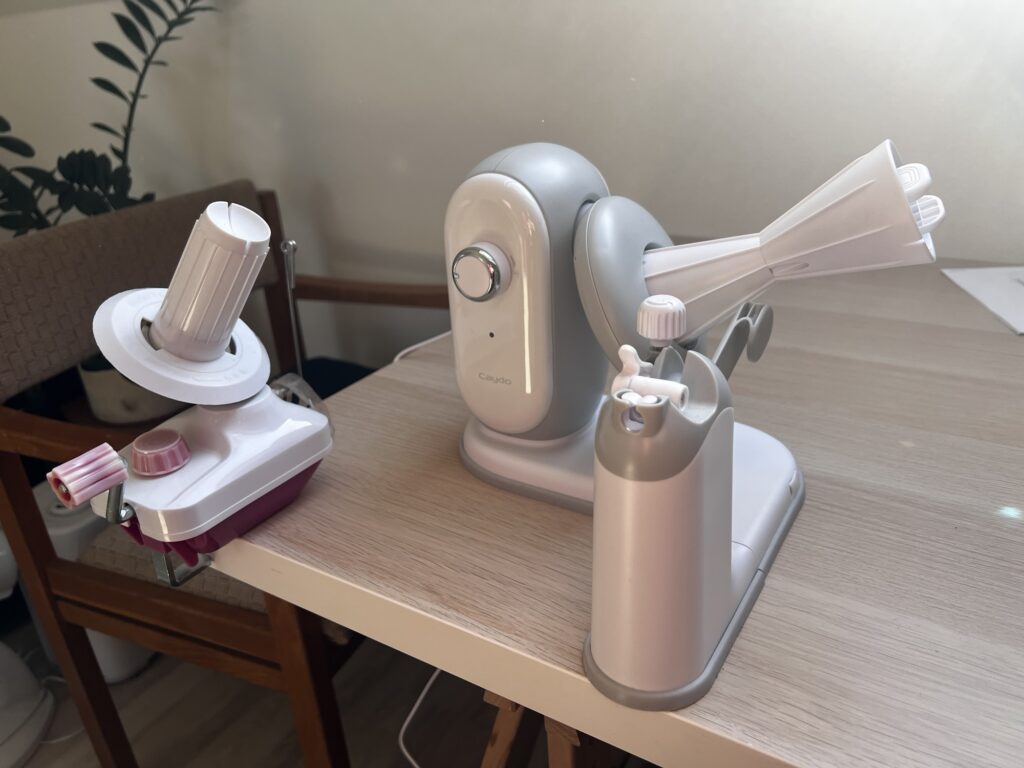 The Caydo A1 Pro Max electric yarn winder on a table beside a small white hand crank yarn winder, showing the electric one takes up about twice the footprint