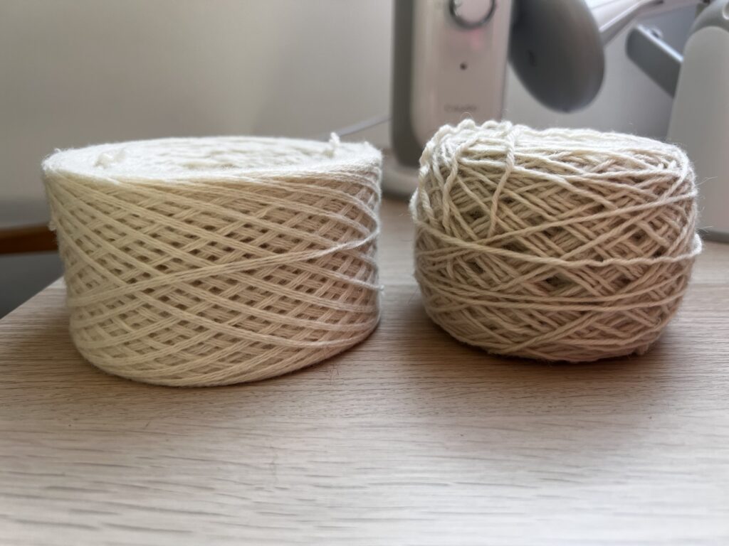 Two cream yarn cakes side by side, the flat sided even cake from the electric winder next to the looser rounder cake from a hand crank winder