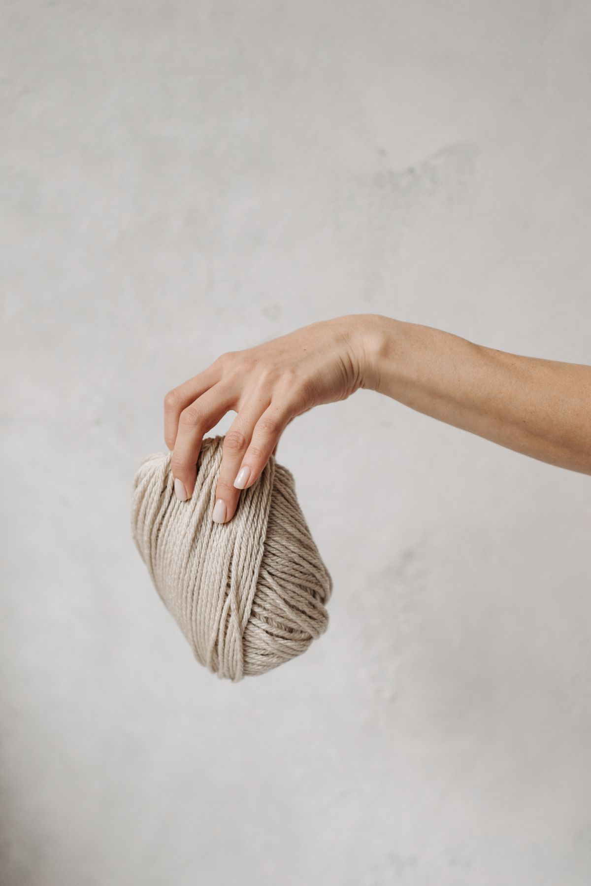 A hand holding a loosely wound ball of undyed natural yarn against a pale wall, the reclaimed yarn relaxed and ready to use